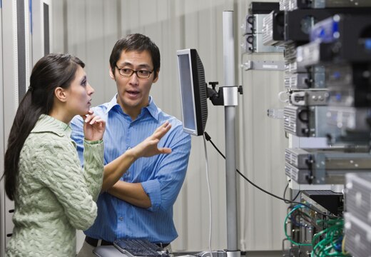 Man talking with a woman standing in front of a monitor in a server room discussing a challenge.